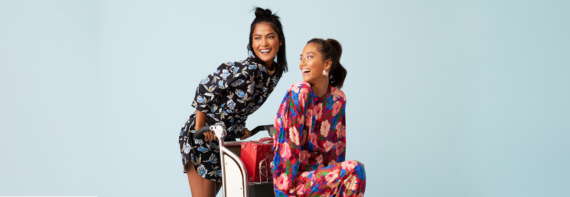 Two young women in bright floral dresses posing on a luggage cart