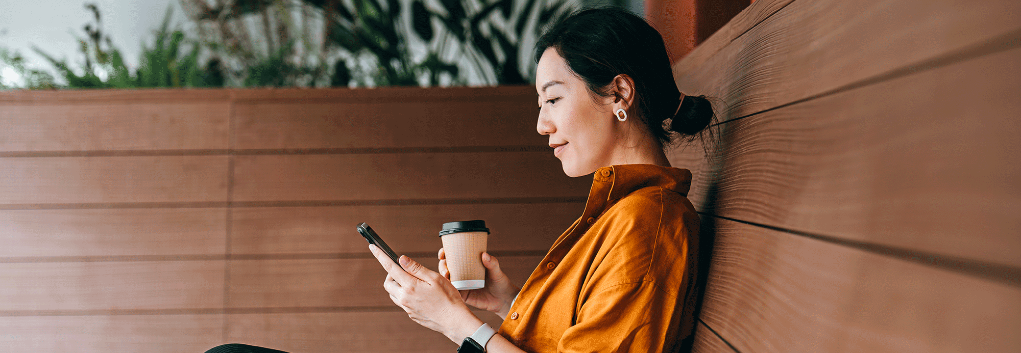 A woman sitting on a bench texting and drinking a coffee