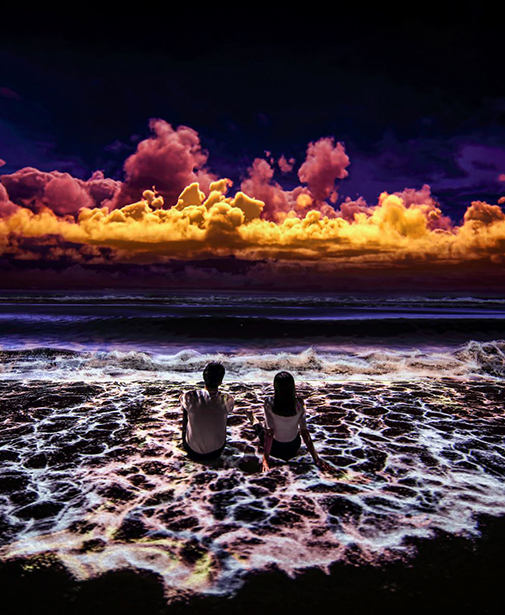 Man and woman sitting on beach, ocean water flowing around them with bright orange and plum color clouds in the sky.