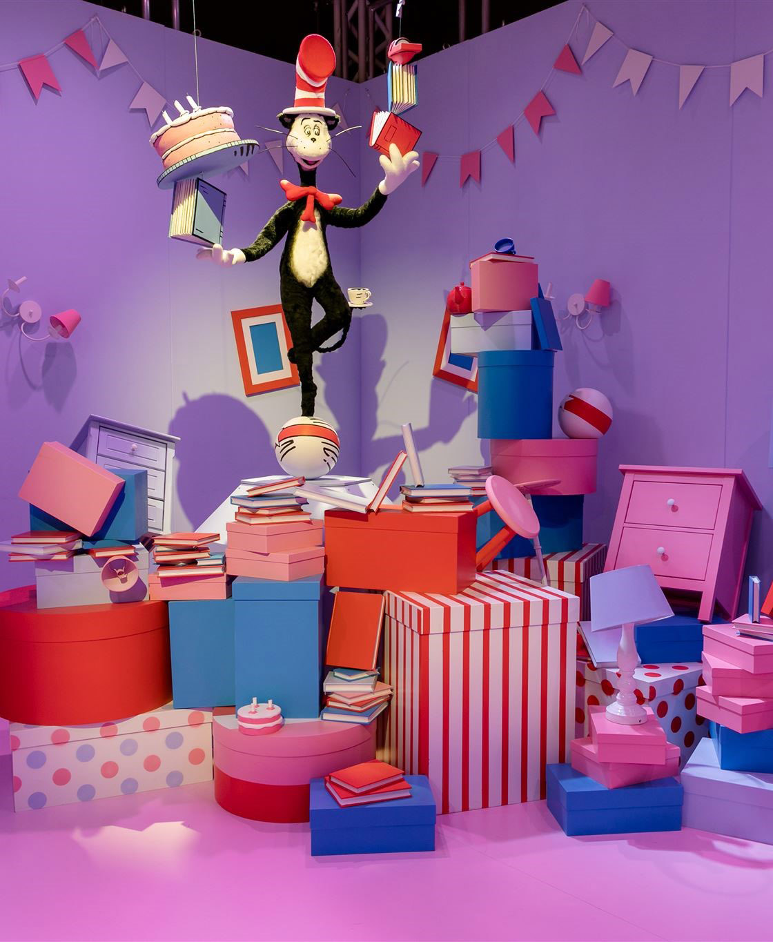 Cat in the Hat holding books and a cake while balancing on a ball stacked on top of colorful books and colorful boxes all stacked in a toppled manner
