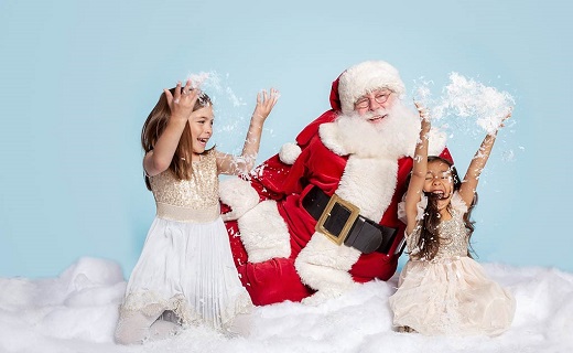 Santa in red suit with white beard and two girls with white dresses playing in snow.