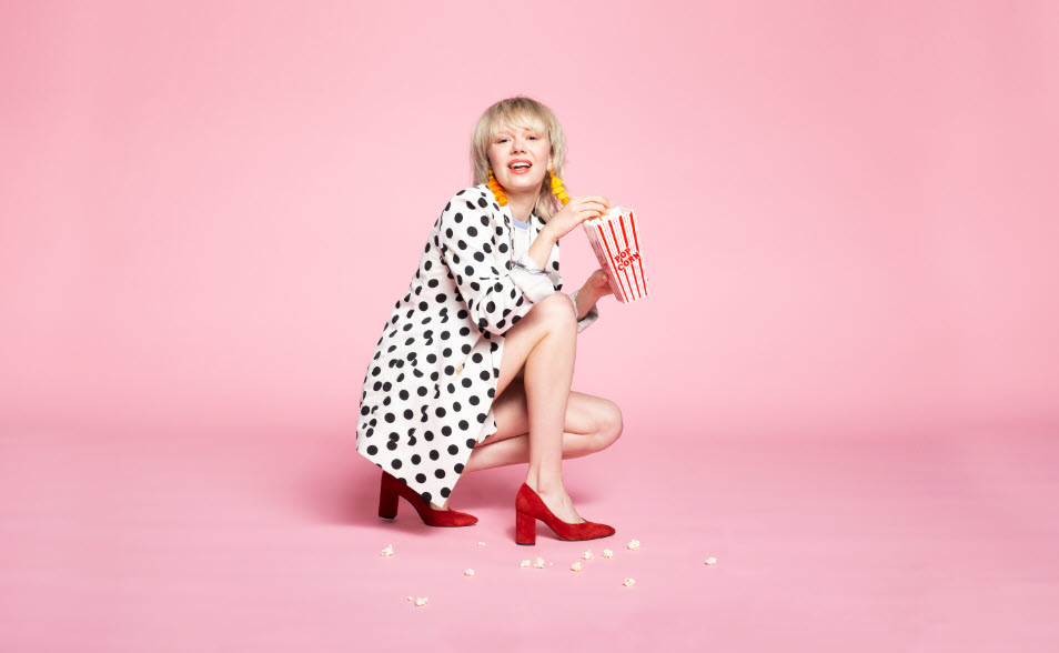 Woman in polka-dots kneeling eating popcorn on pink background