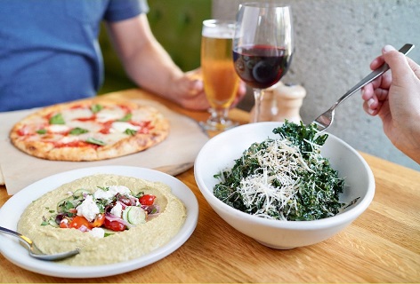 Two people eating True Food Kitchen entrees including humus, a salad and pizza. They are also drinking a glass of wine and a beer.