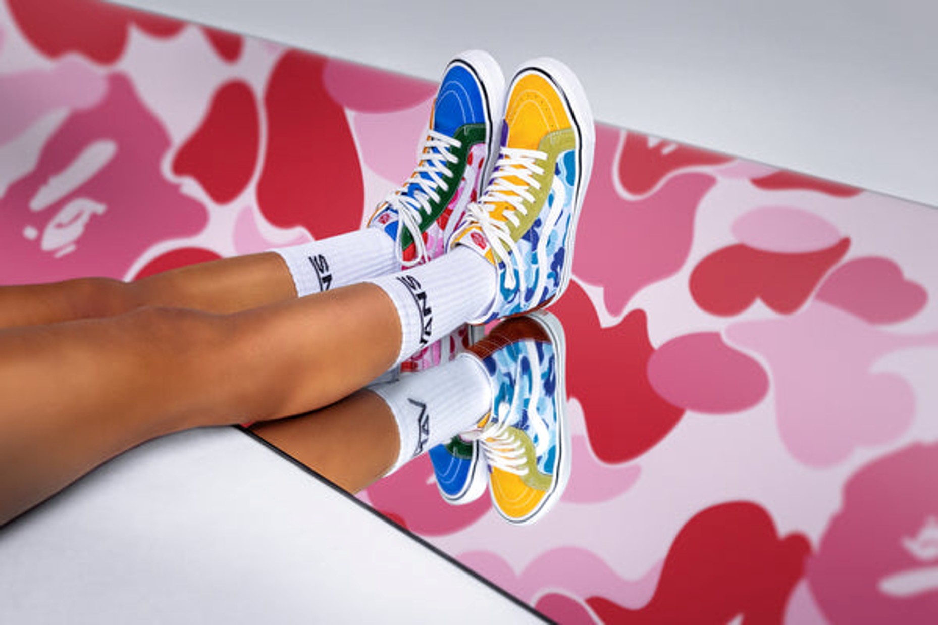 Persons legs over a mirror wearing white vans socks and rainbow colored vans x BAPE shoes. 