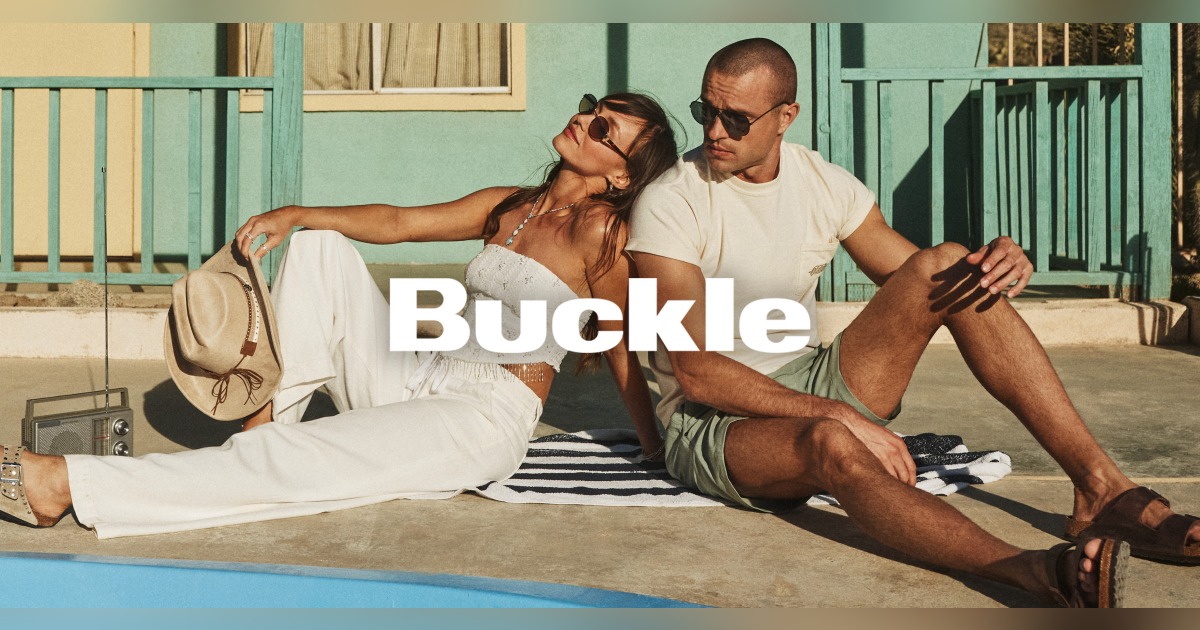 A couple sitting on a beach towel leaning on each other. The Buckle logo is over the top of the image. 