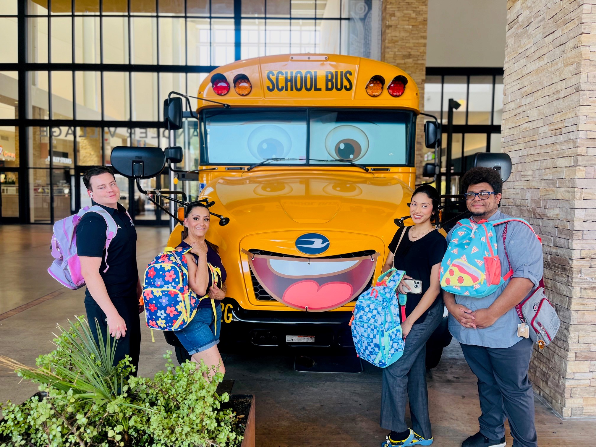 School bus with a cartoon face on the front of the bus that looks like a smiley face. Four people in the front donating back packs with school supplies.