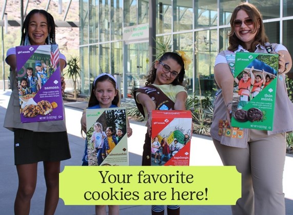 Three little girls dressed with brownies and girl scout sashes holding a box of girl scout cookies with a troop mom holding a box of cookies. 