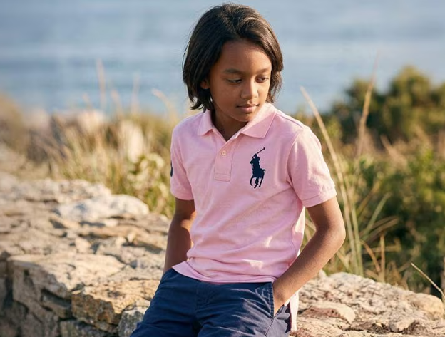 Young boy in pink polo shirt and navy shorts sitting on a brick fence at the ocean 