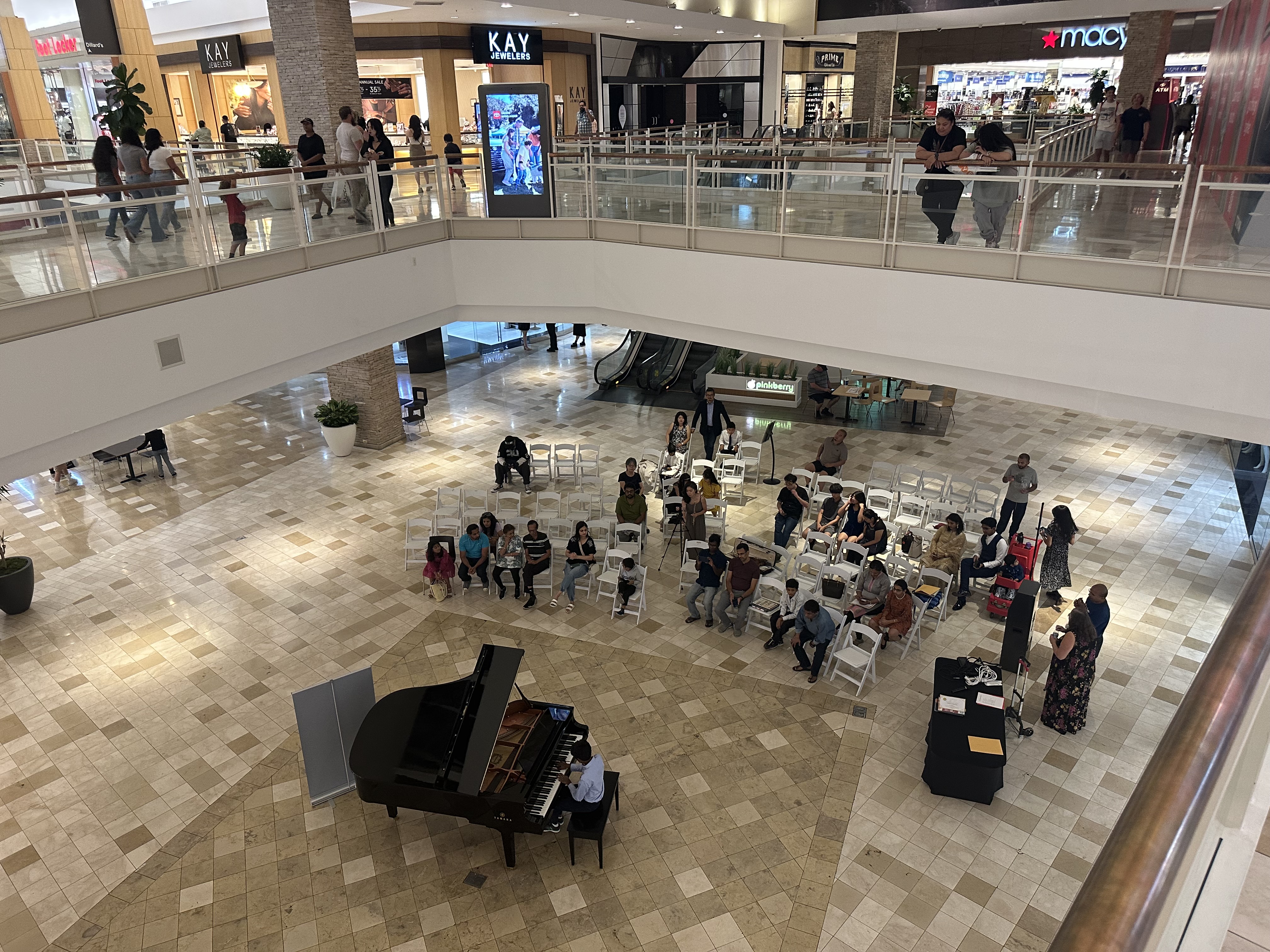 Image of a grand piano in a shopping mall with people listing to students play.
