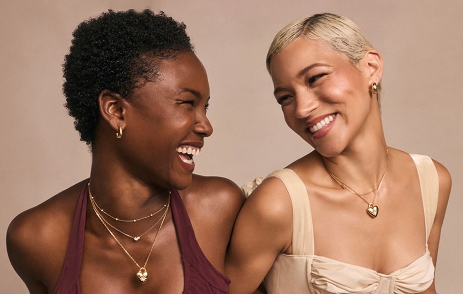 Two ladies smiling at each other wearing heart shaped kendra scott gold necklaces 