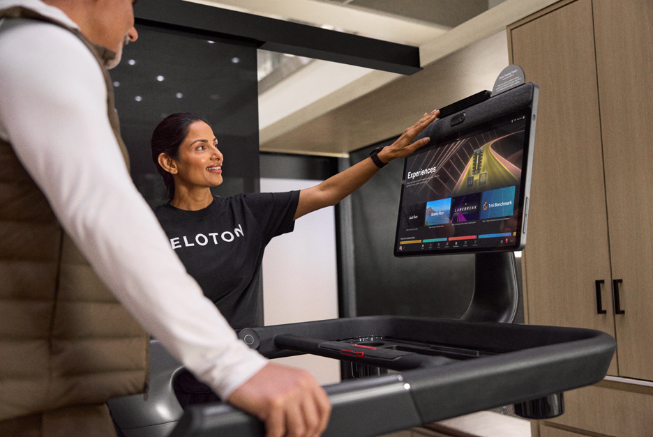 Lady wearing a Peloton shirt showing a man on a treadmill how the touch screen works. 