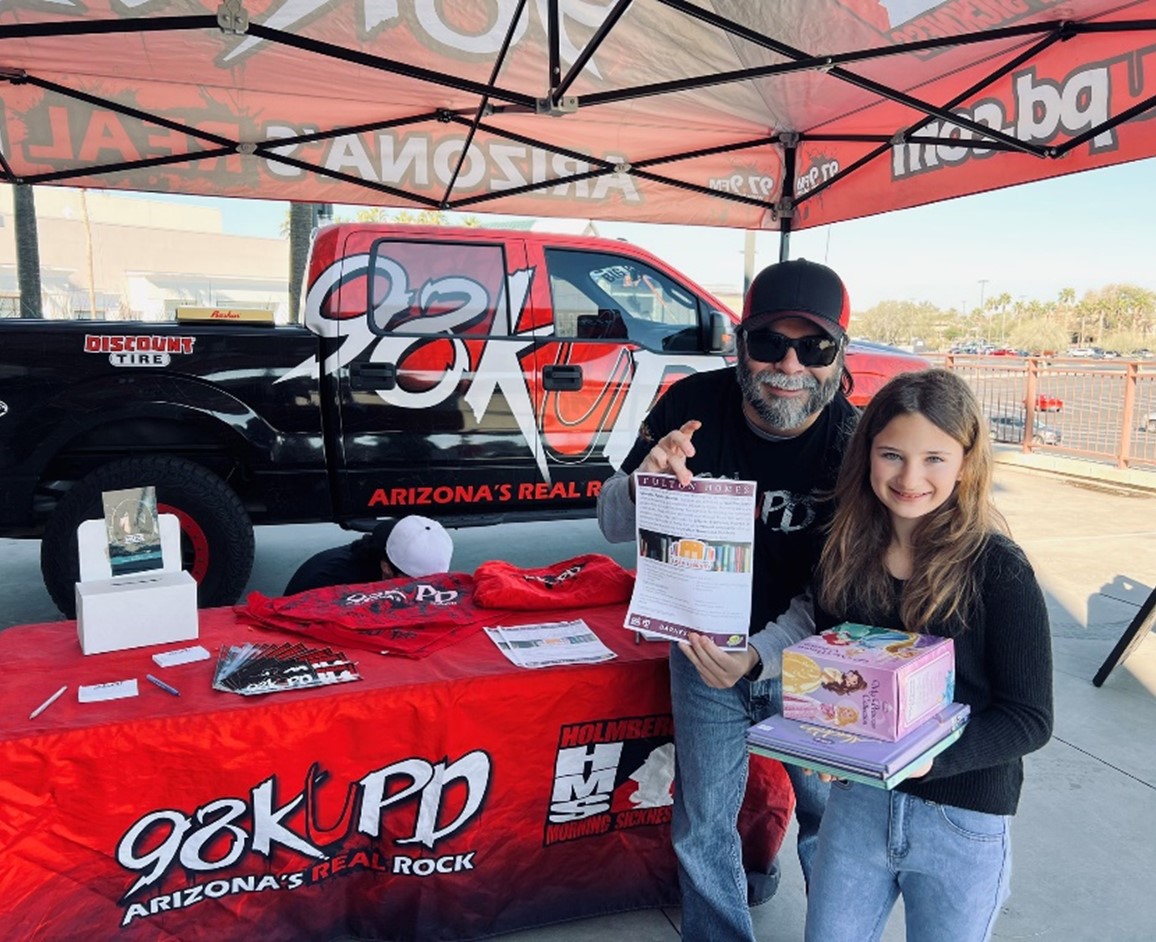 Radio Dj and a child holding books to donate, standing in front of a table that says 98KUPD Arizona's Real Rock. 