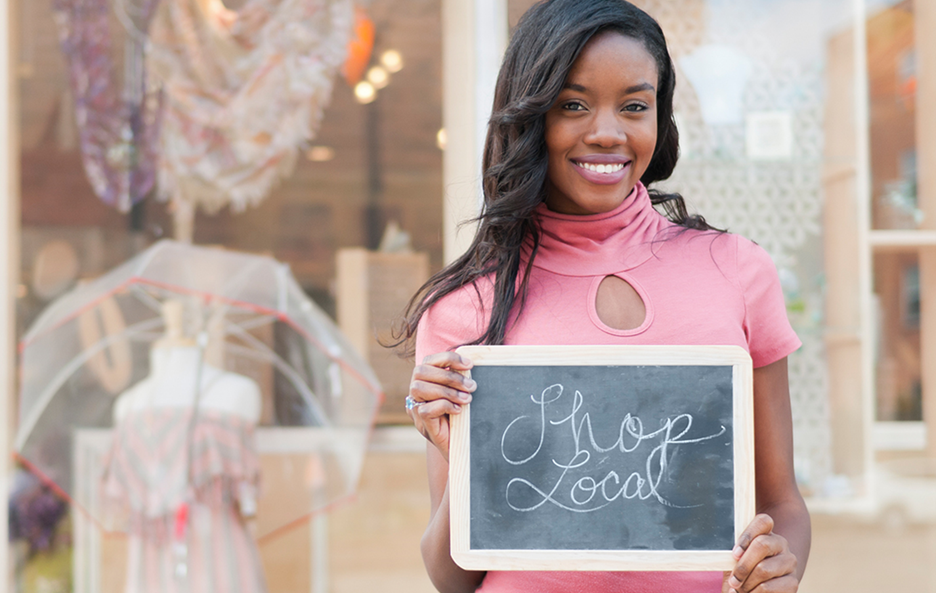 A woman in a pink top holding a small chalk board that says "Shop Local". 