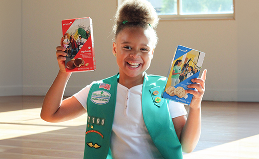 Little girl in a girl scout vest holding up two boxes of cookies. 