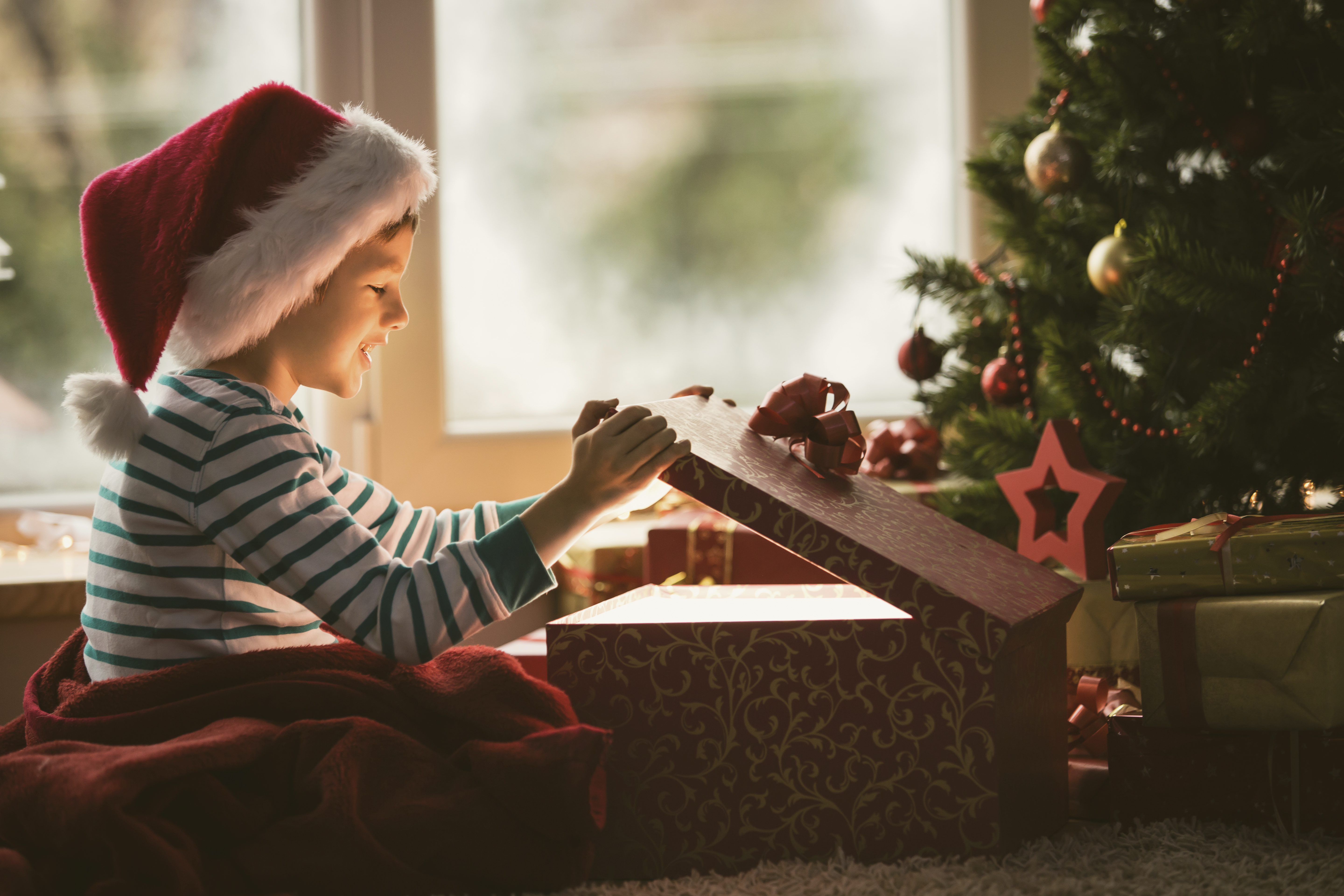 boy in pajamas and a Santa hat opening a gift box in front of the Christmas tree. 