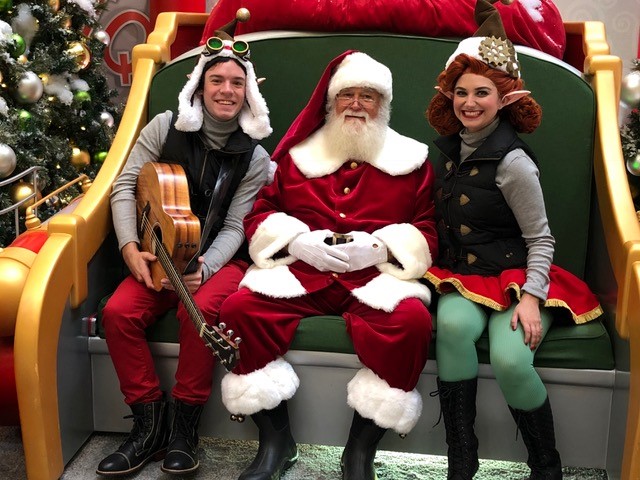 two elves sitting with Santa on his big rocket chair