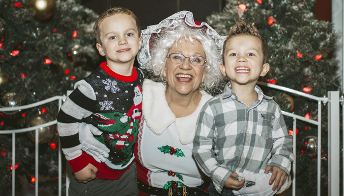 Mrs. Claus hugging two young children in front of Christmas Trees