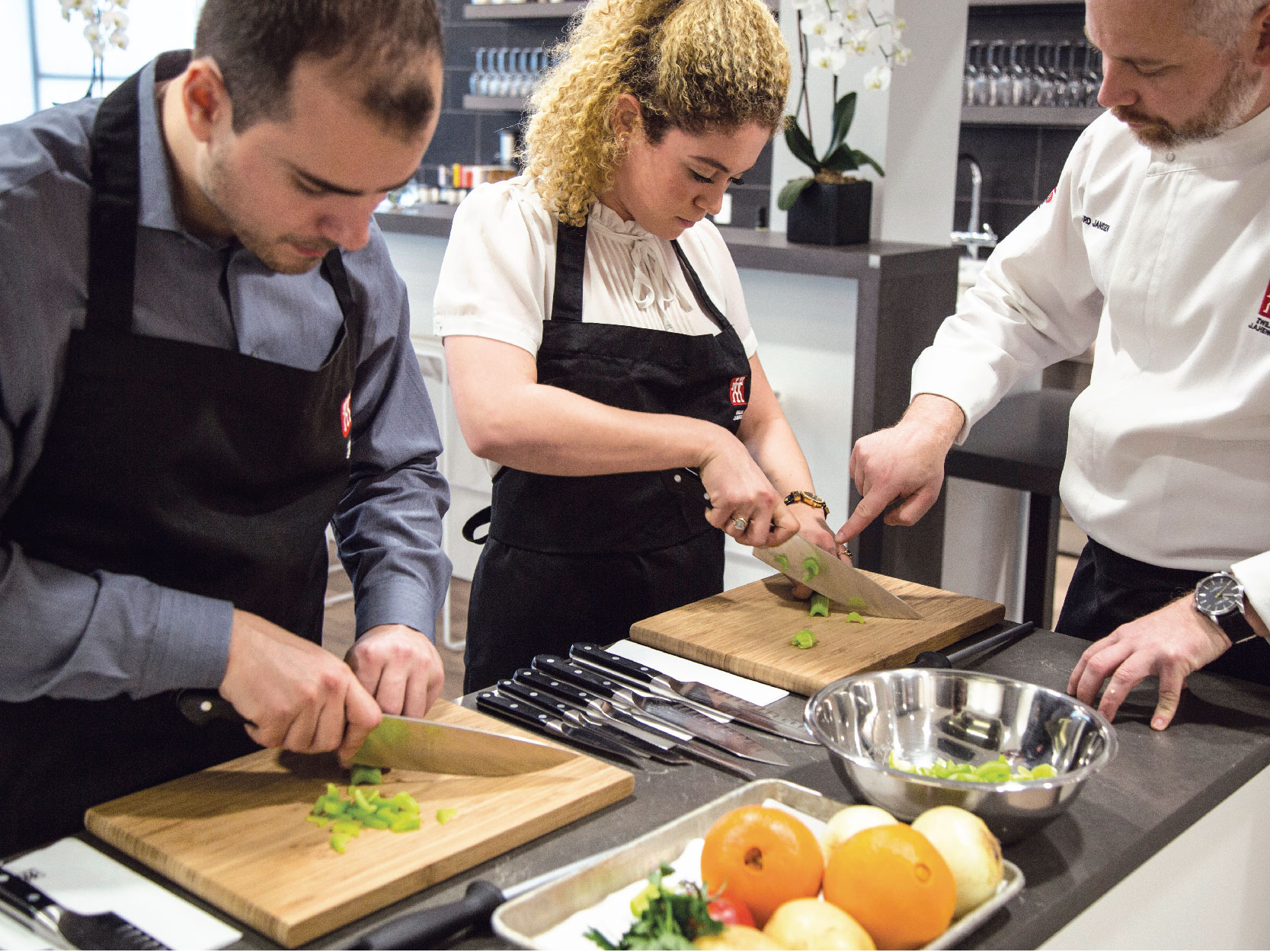 Two students, wearing black aprons, being taught knife cutting techniques by a Zwilling employee. 