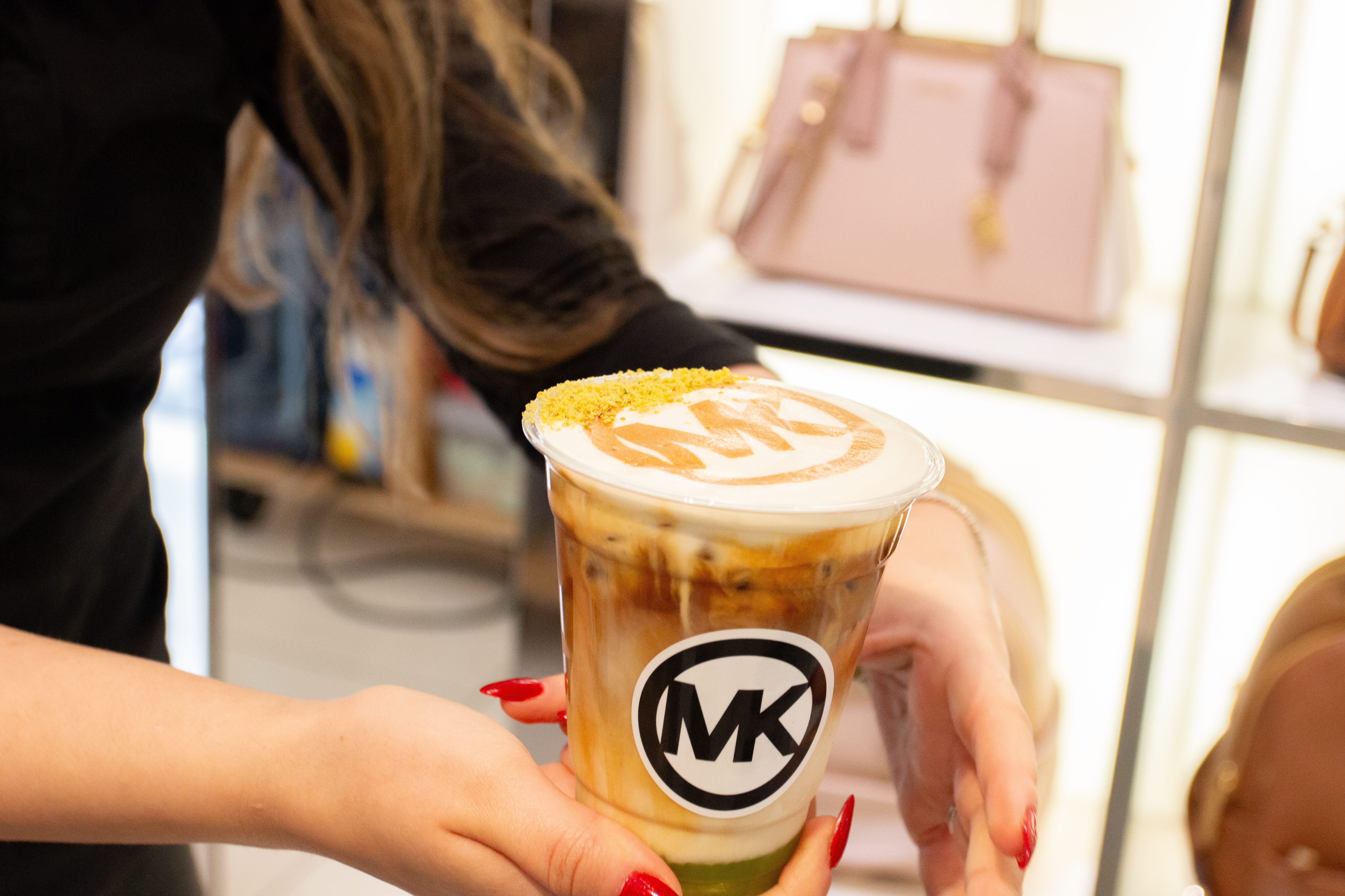 A close up images of a coffer in the hands of a women.  The plastic coffee cup has the Michael Kors MK Logo on the cup and also designed on the top of the beverage. 