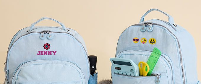 Two light blue backpacks with monogramming. The backpack on the left is monogrammed with a red flower and the name Jenny in purple. The backpack on the right has three emojis in a row: a smile face with heart sunglasses, sticking out its tongue and a smiley face.