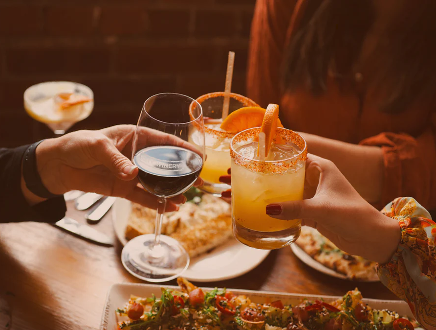 Warm, golden-lit photo of hands toasting with a City Winery red wine glass and citrus cocktails with salted rims, over plates of shared food