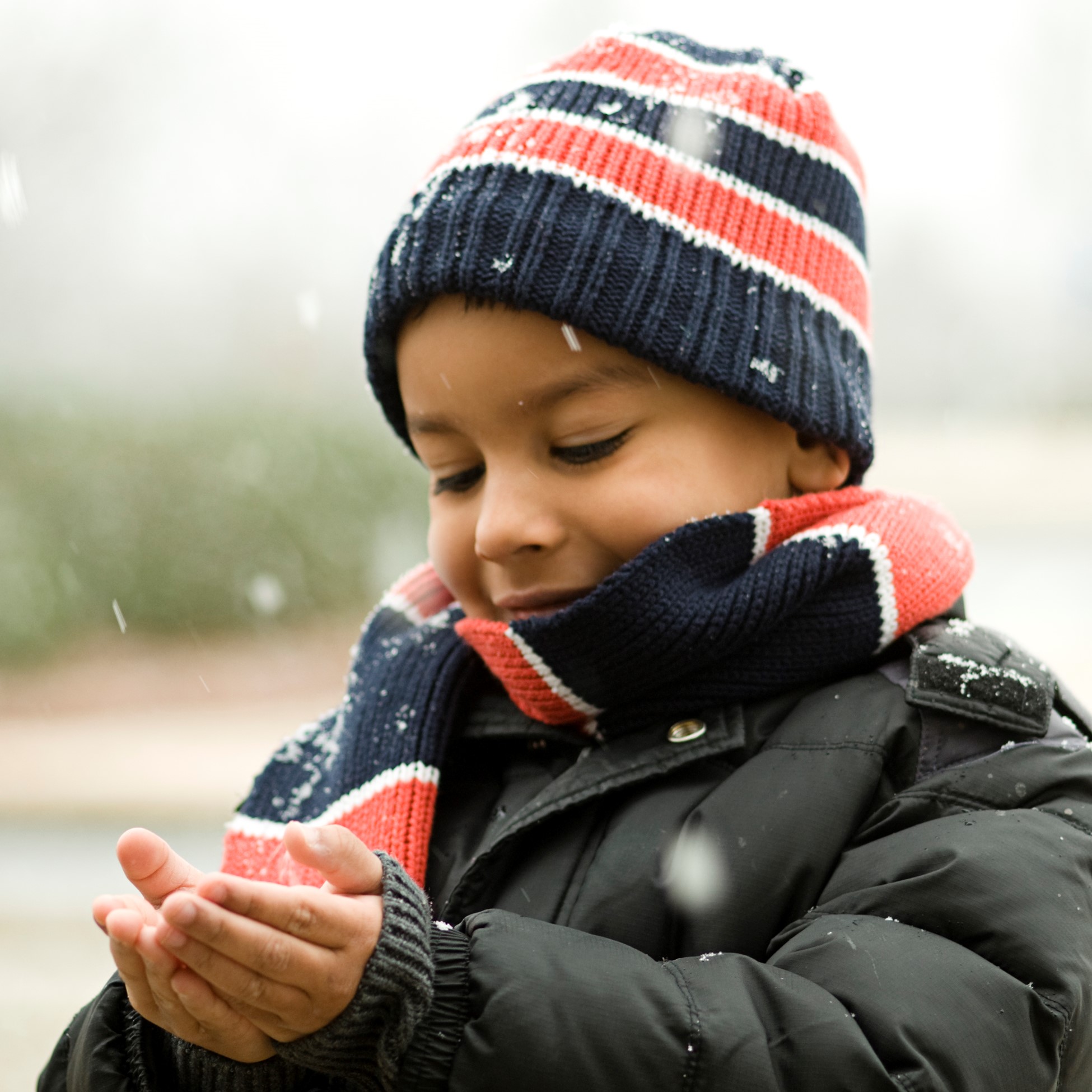 Little boy in winter coat, scarf and hat catching snow in his hands. 