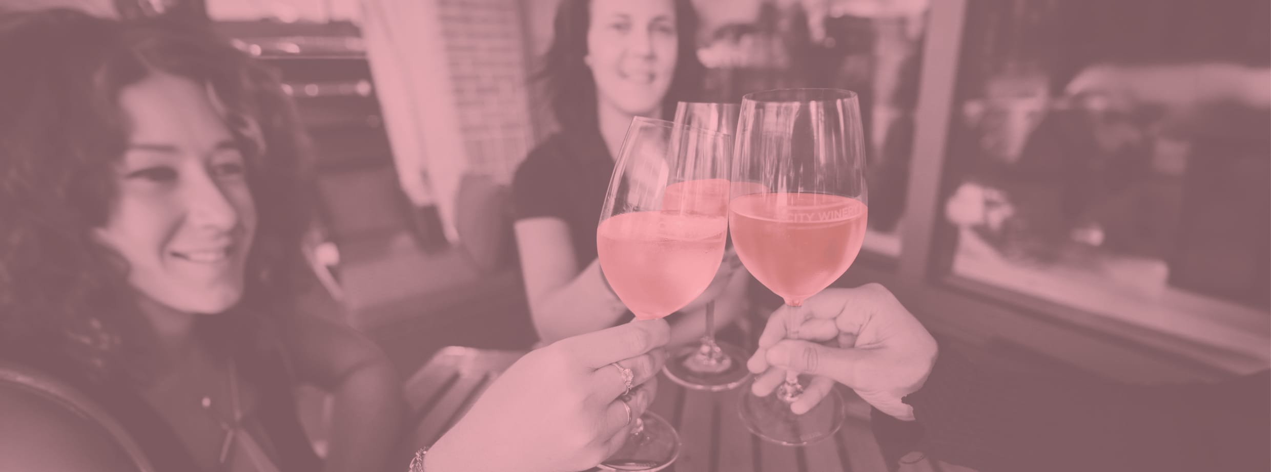 Photo with a pink duotone filter of two smiling women toasting with glasses of rosé wine at an indoor gathering