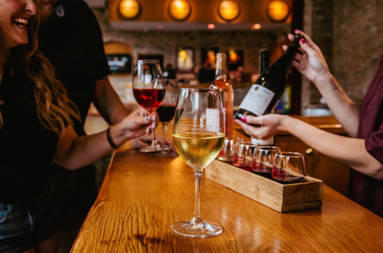 Guests enjoying red and white wine glasses and a wine flight at a City Winery bar, with a bottle being poured in the background