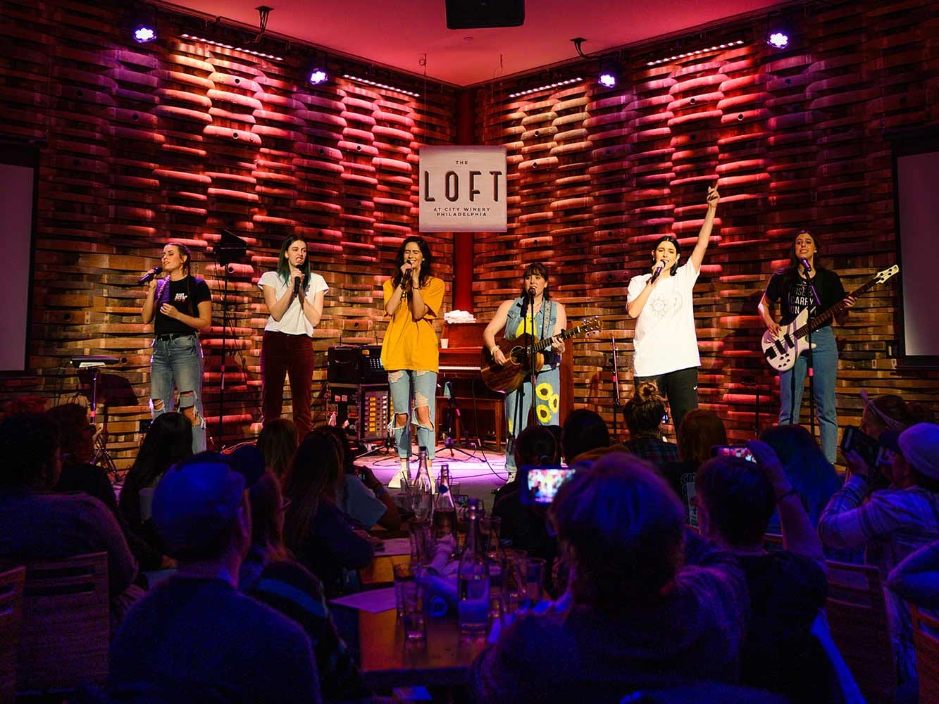 A group of musicians performing on stage at The Loft at City Winery Philadelphia, in front of a wine barrel backdrop with a crowd watching under red stage light