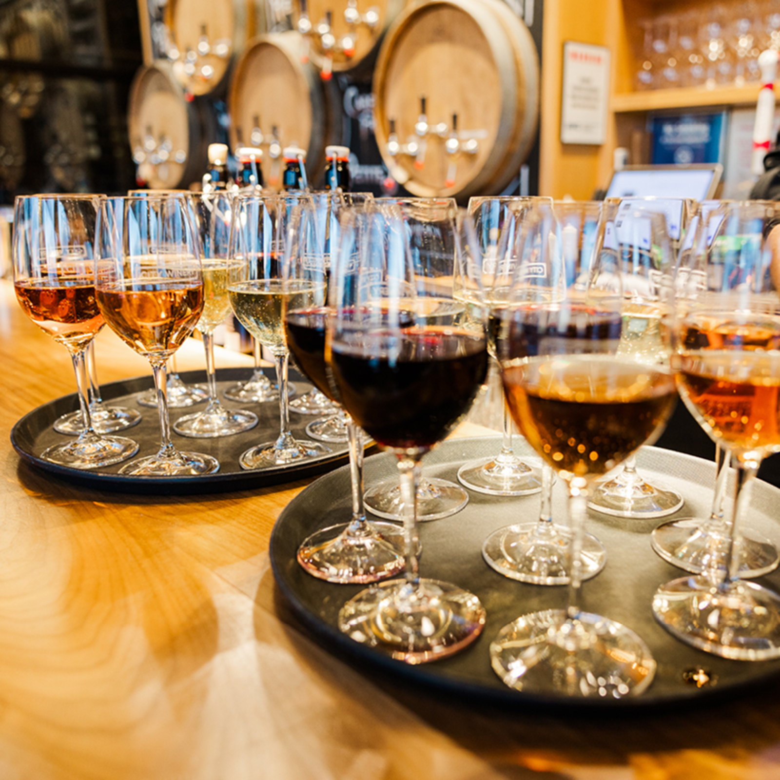 Two round trays of assorted red, white, and rosé wine glasses on a wooden bar, with wine barrels stacked in the background