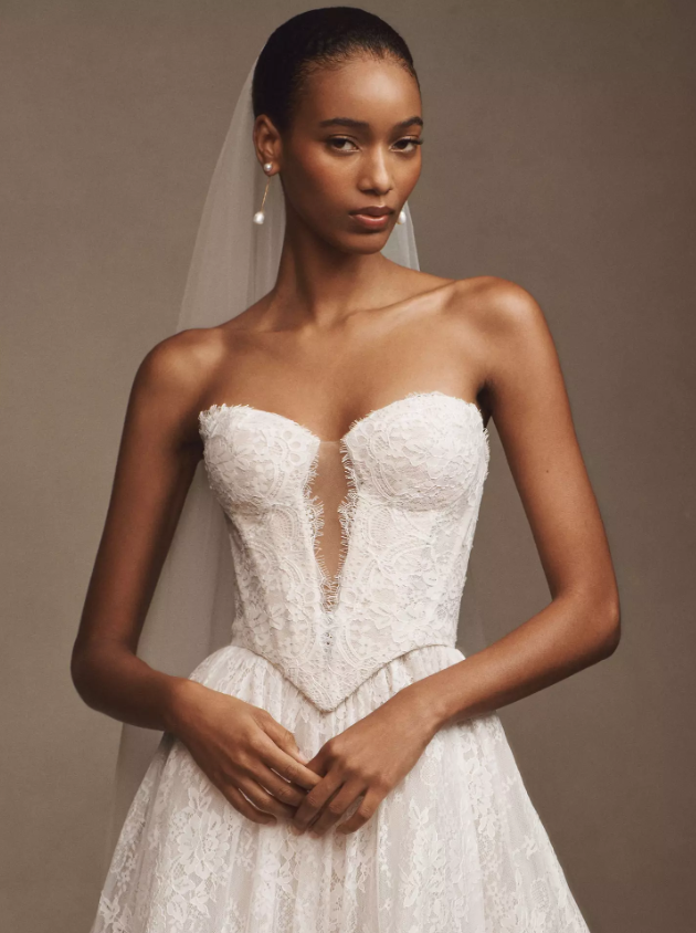 A model wearing a strapless lace wedding gown featuring a deep illusion neckline, fitted bodice, and full A-line skirt, paired with a simple veil and pearl earring.
