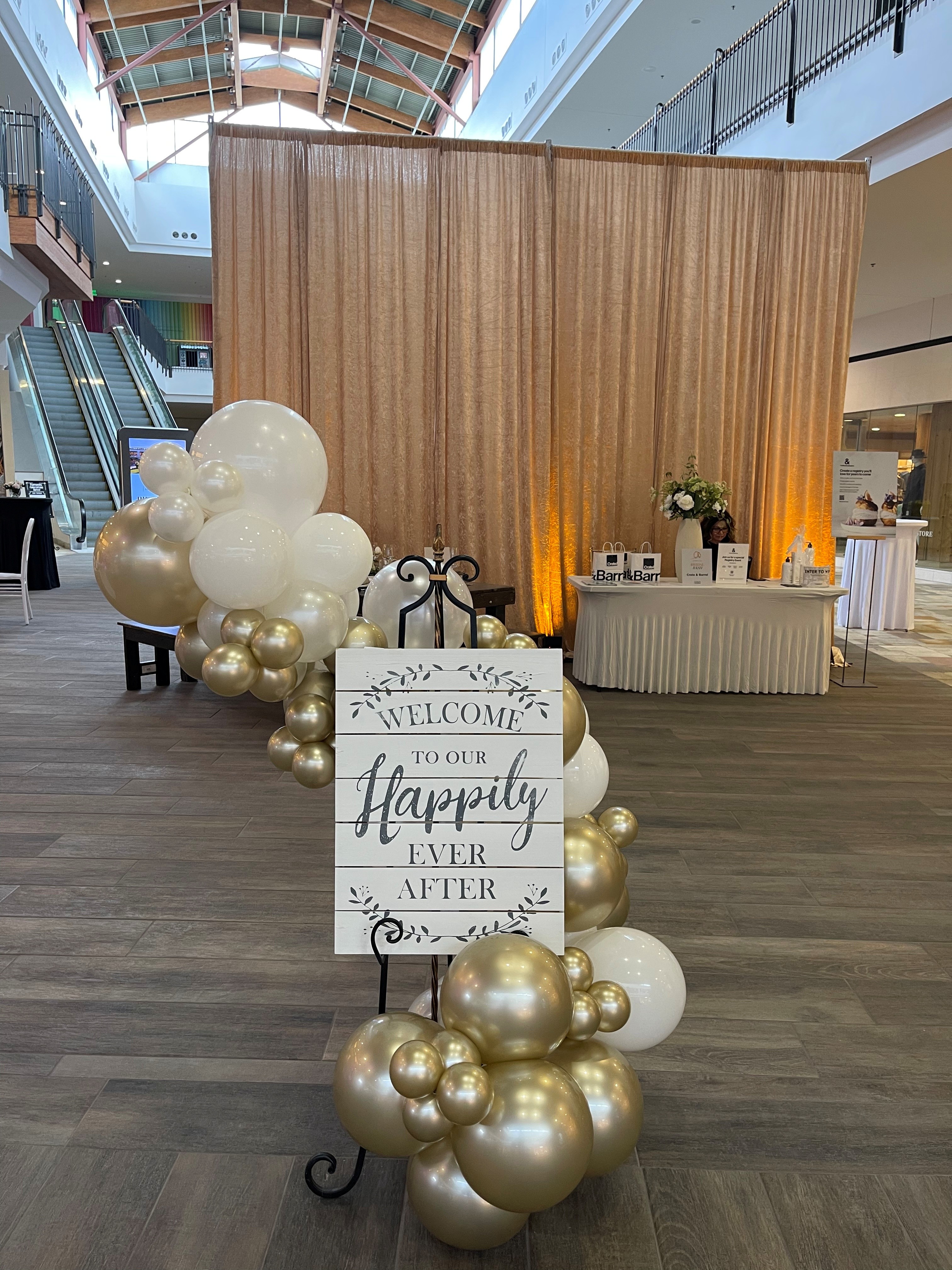 Wedding-themed welcome display featuring white and gold balloon arrangements and a sign reading “Welcome to Our Happily Ever After” in a mall event space.