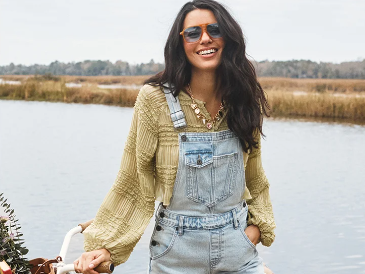 A person standing outdoors near a calm body of water, wearing light denim overalls layered over a textured long-sleeve top, with one hand resting on a bicycle.