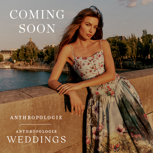 Woman leaning on a stone wall wearing a floral dress. Body of water and buildings in the background. Text reads "Anthropologie. Anthropologie Weddings."