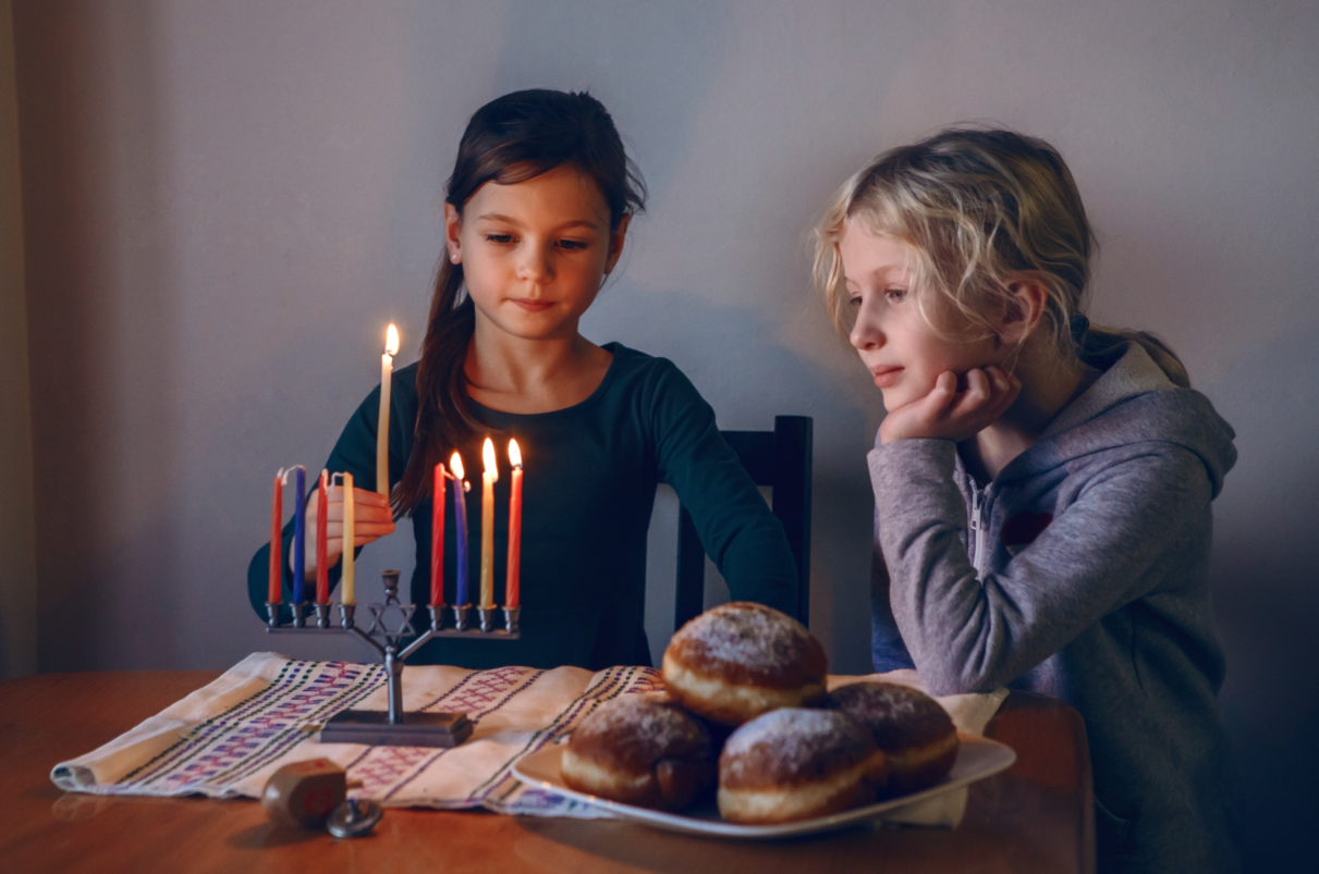 Two young girls lighting candles and standing by a table with a plate full of donuts.