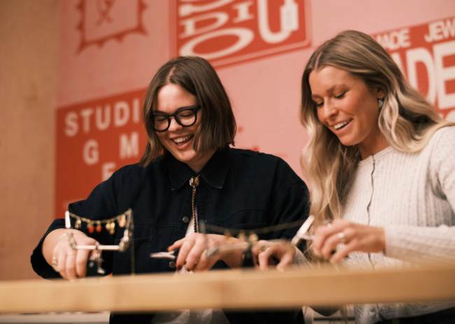 Two people seated at a worktable participate in a jewelry-making workshop, using tools and materials to create custom pieces, with Gem Studio branding visible in the background.