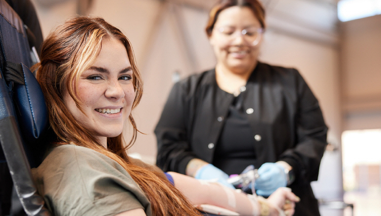 A smiling woman donates blood while a healthcare professional prepares the donation in a clinic setting.
