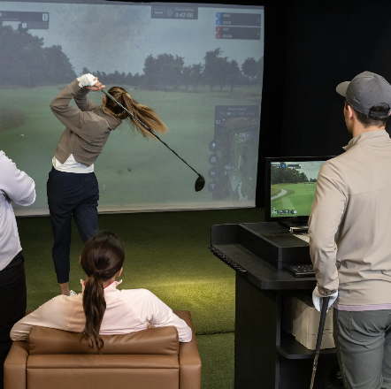 Woman, with a Golf Club playing golf in a golf simulator - while several people watch. 