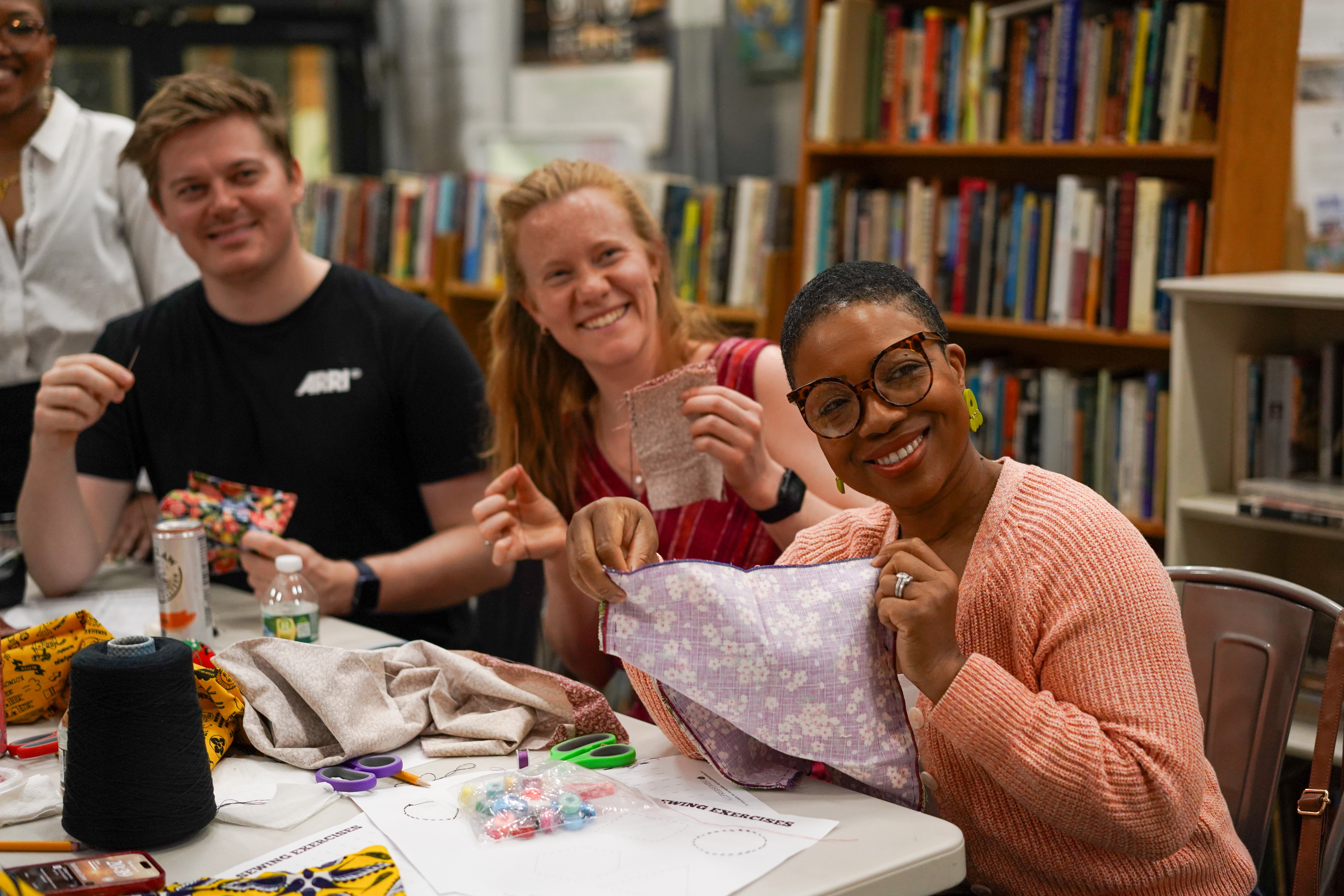 three smiling people sitting at table covered with sewing and craft accessories, holding up fabric and material. 