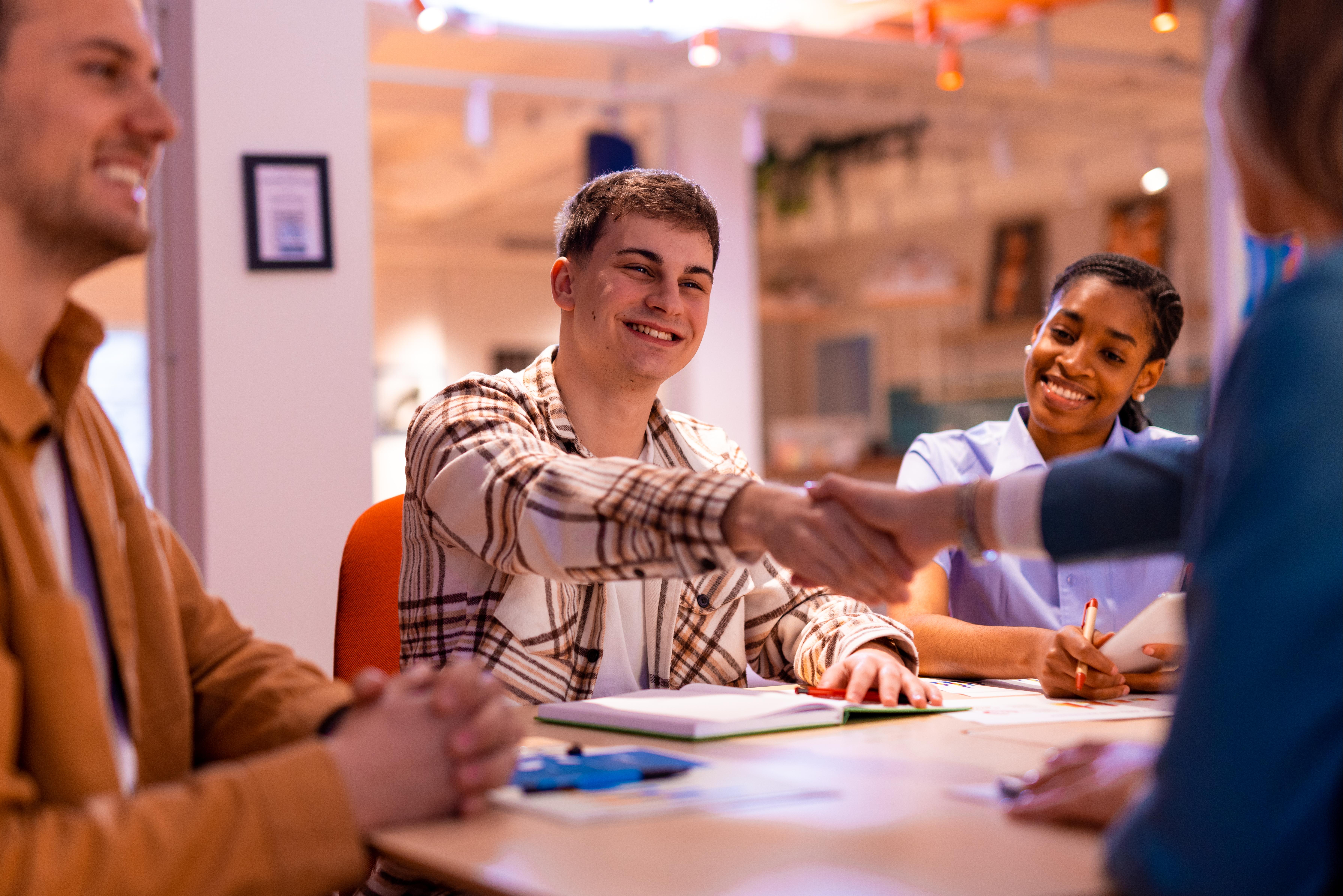 Four coworkers smiling at a table during a meeting; two shake hands while others take notes. 