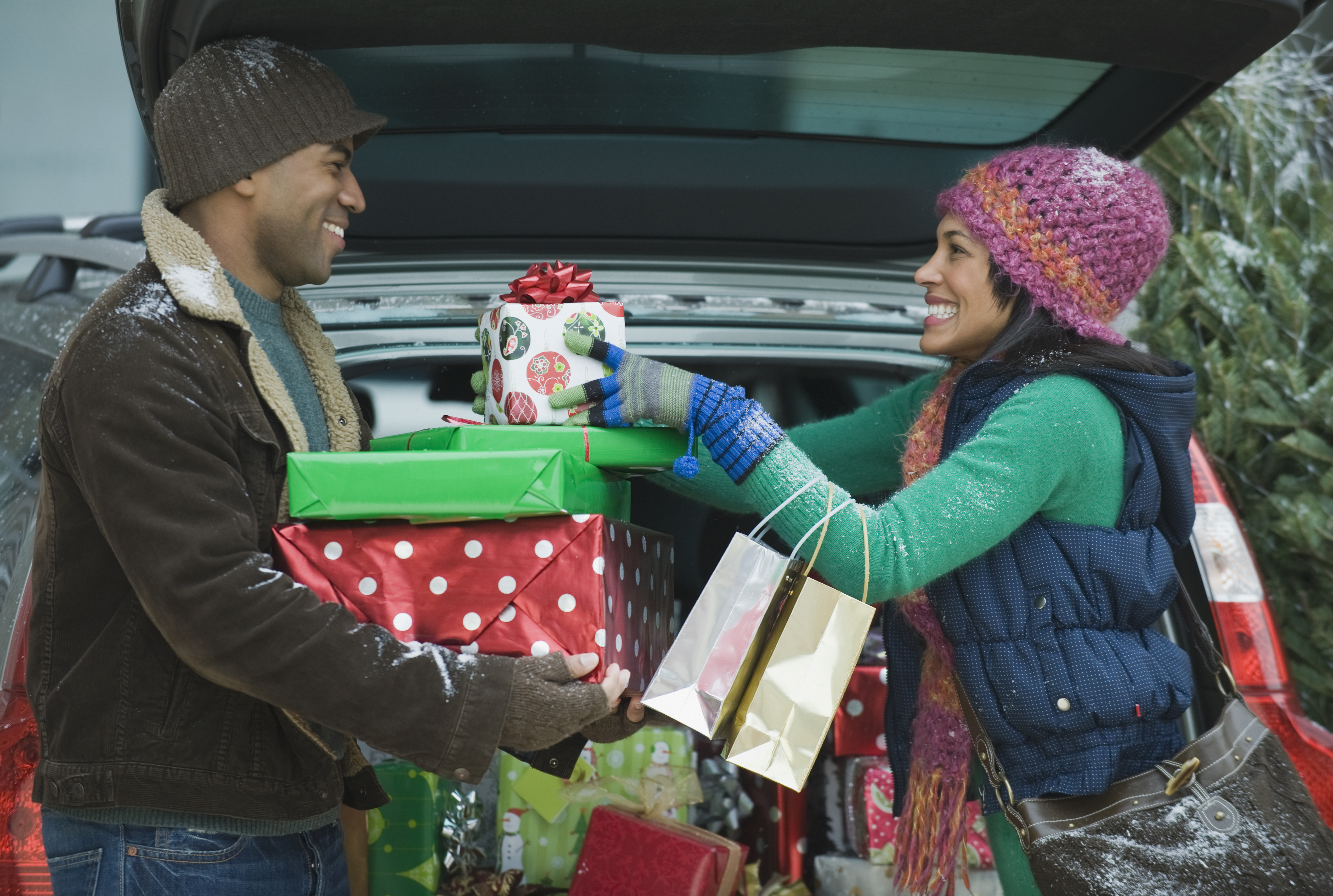 a couple looking at each other and smiling while  in front of a car with many wrapped presents in hand
