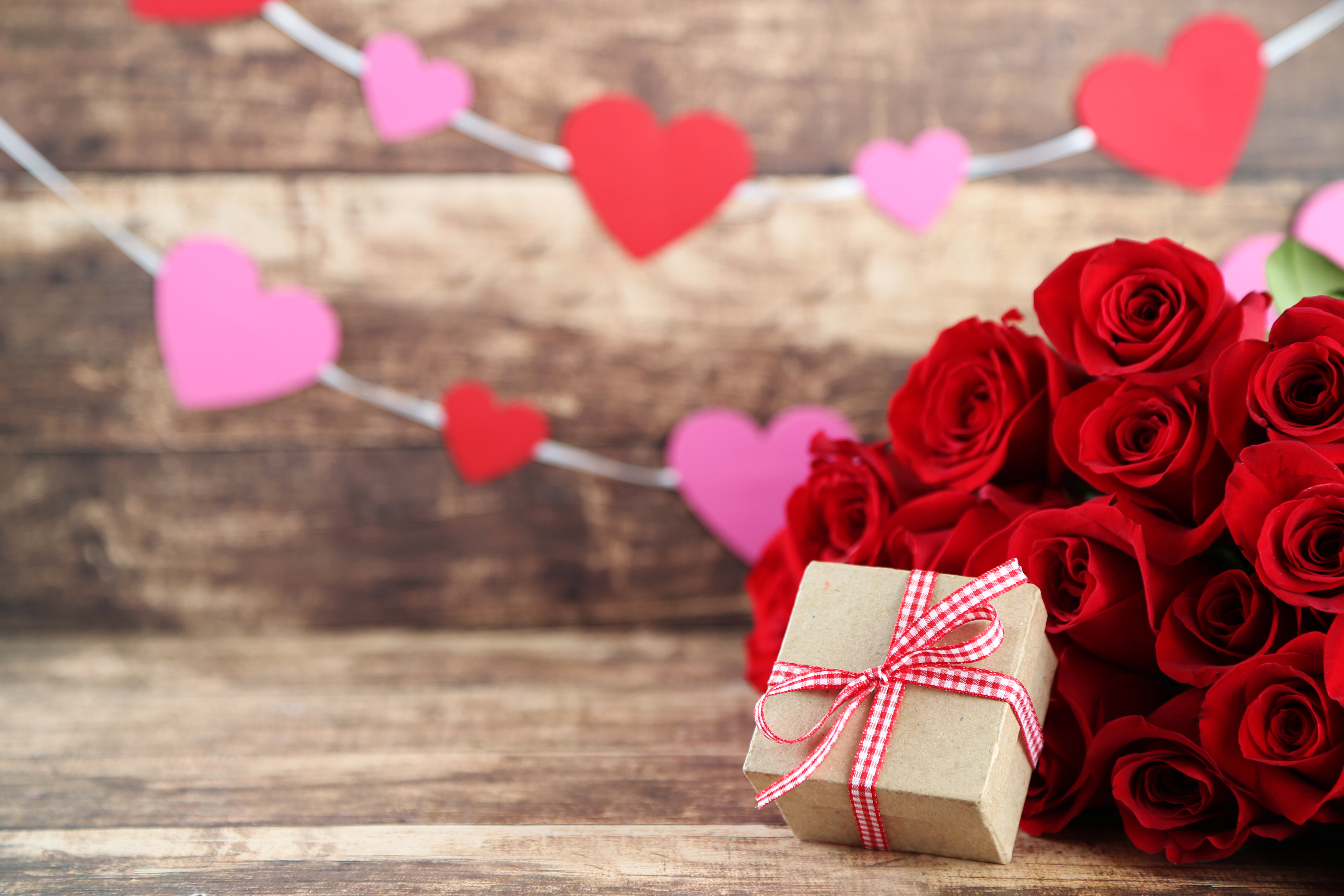 wood background with a pink and red heart garland and a bouquet of red roses with a small box with a bow in front of the bouquet. 
