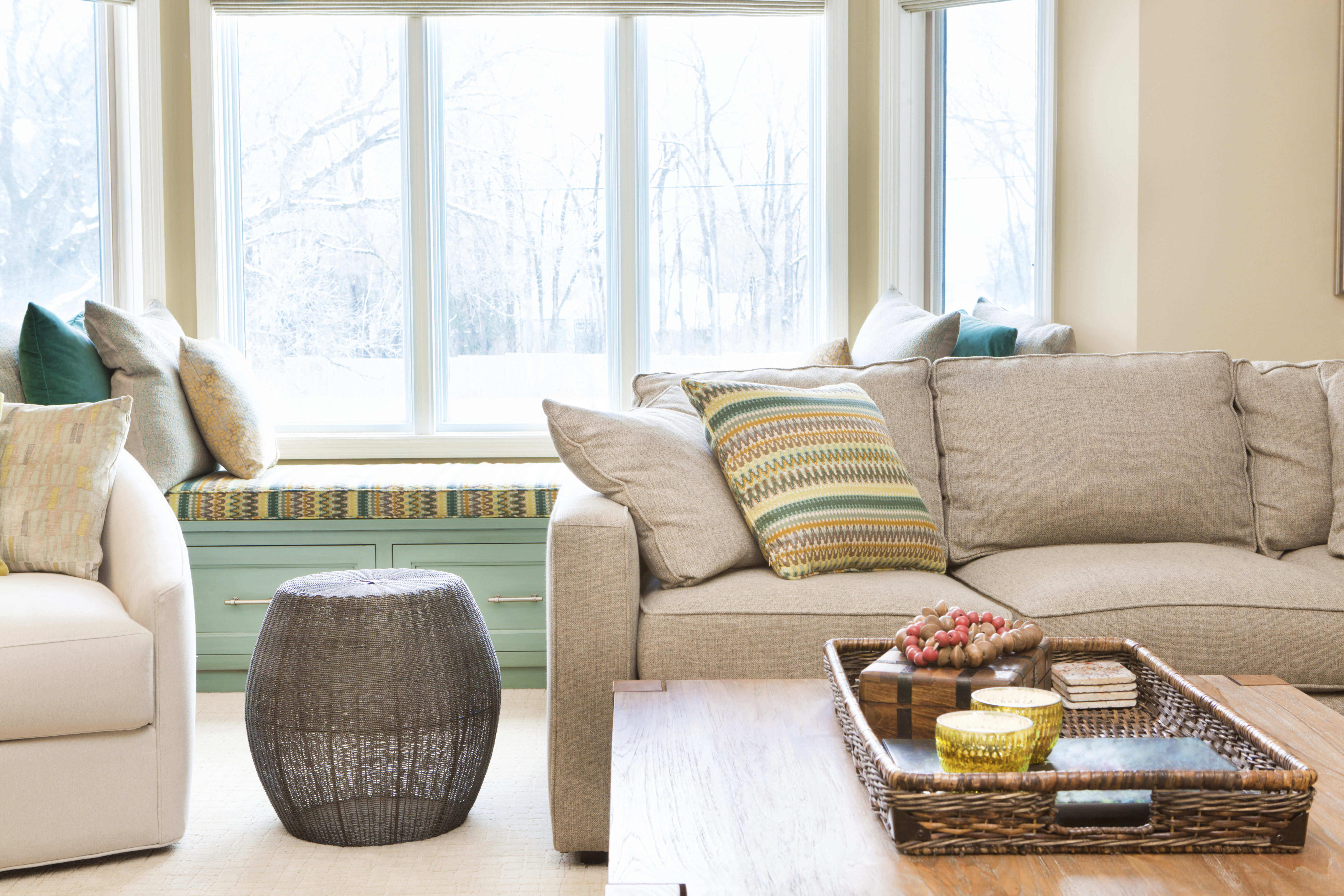 living room with beige colored couch and coffee table, window in the background with trees and a lawn, a window seat with pillows in front of the window