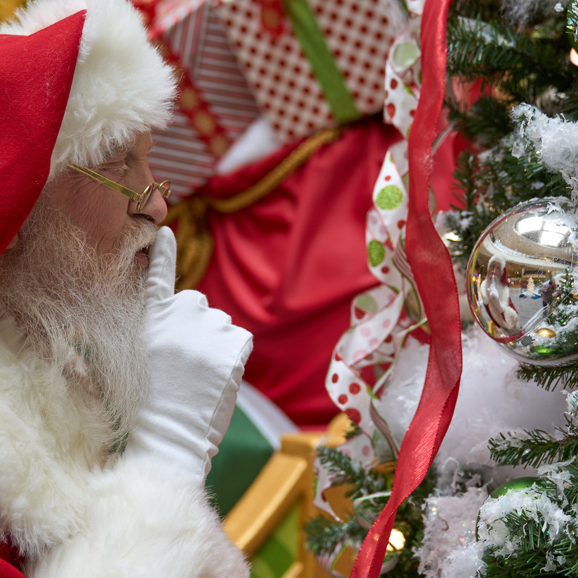 Santa Looking at His Reflection in a Silver Ornament While He Makes the "Be Quiet" SIgn with His Hand to His Mouth