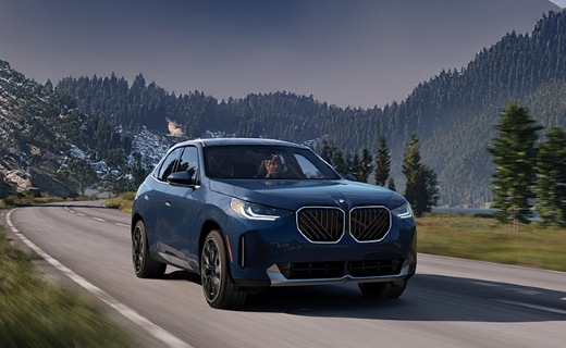 A blue BMW SUV is driving on a scenic mountain road surrounded by trees and hills under a clear sky.