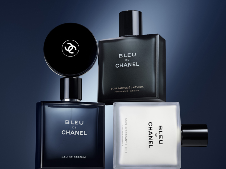 Three Bleu de Chanel grooming products, including Eau de Parfum, hair care, and moisturizer, displayed against a dark blue background.