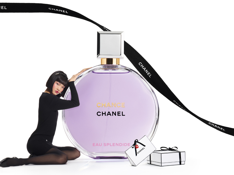 Model posing beside oversized Chanel Chance Eau Splendide perfume bottle with gift boxes.