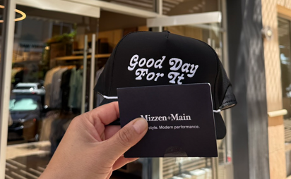 Hand holding a “Mizzen+Main” card and a black cap that reads “Good Day For It” in front of a clothing store.