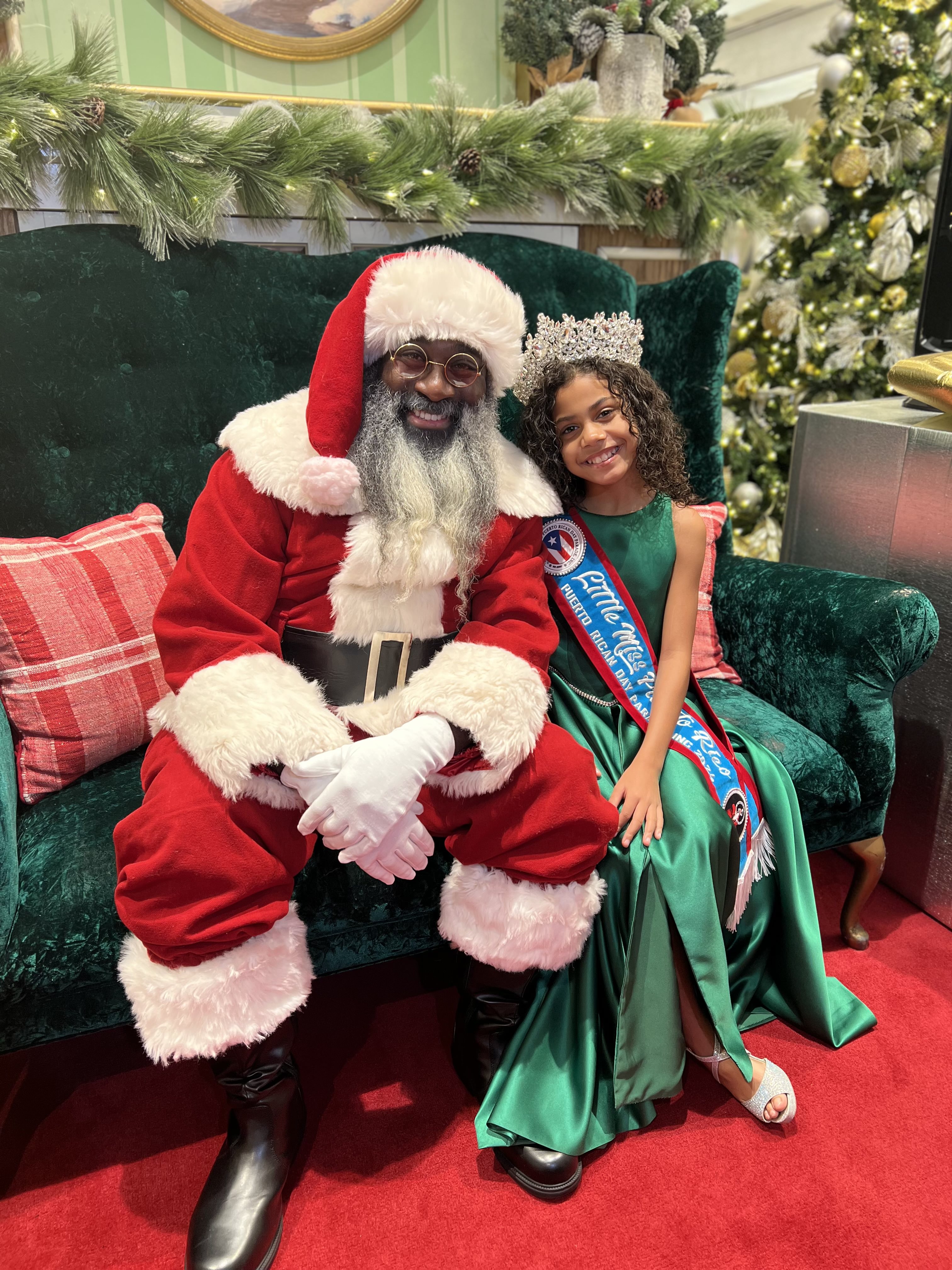An African-American Santa Claus sitting next two a little girl with a green dress and tiara. 