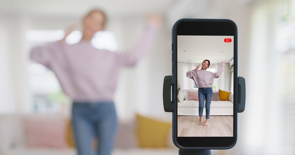 Woman in pink top and denim pants apparently filming herself dancing with a cell phone on a stand.